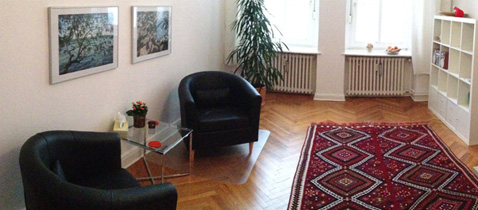Freundliches und behagliches Zimmer mit zwei Sesseln an 
        	einem Glastisch, zwei Bildern an der Wand und einer Pflanze am Fenster.